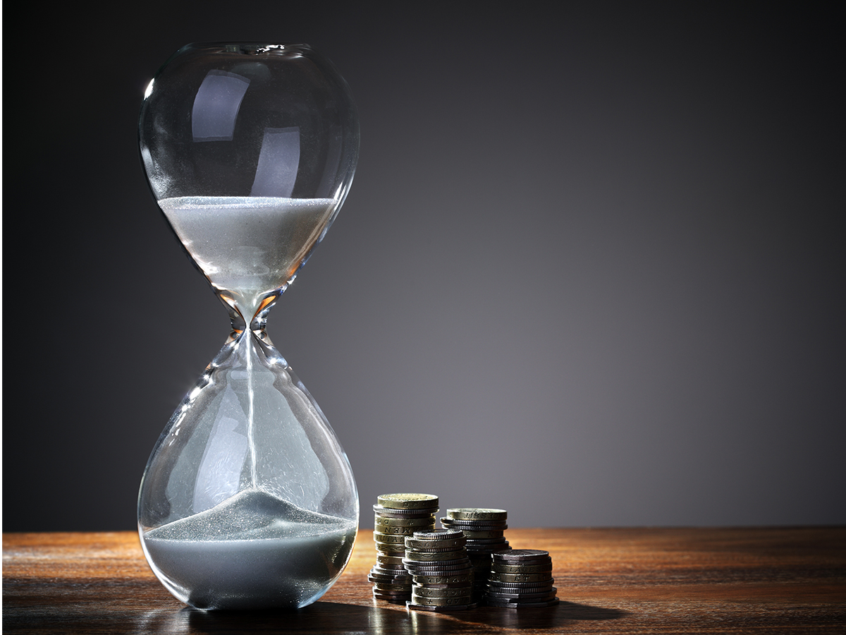 An hourglass pouring with a stack of silver coins next to it.